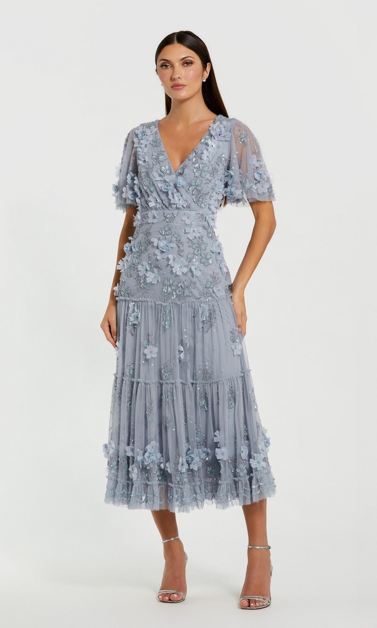 A woman with long dark hair wears the Mac Duggal 9207 A-Line Wedding Guest Dress, featuring a light blue floral design, short sleeves, V-neckline, tiered tea-length skirt, and paired with strappy heels.
